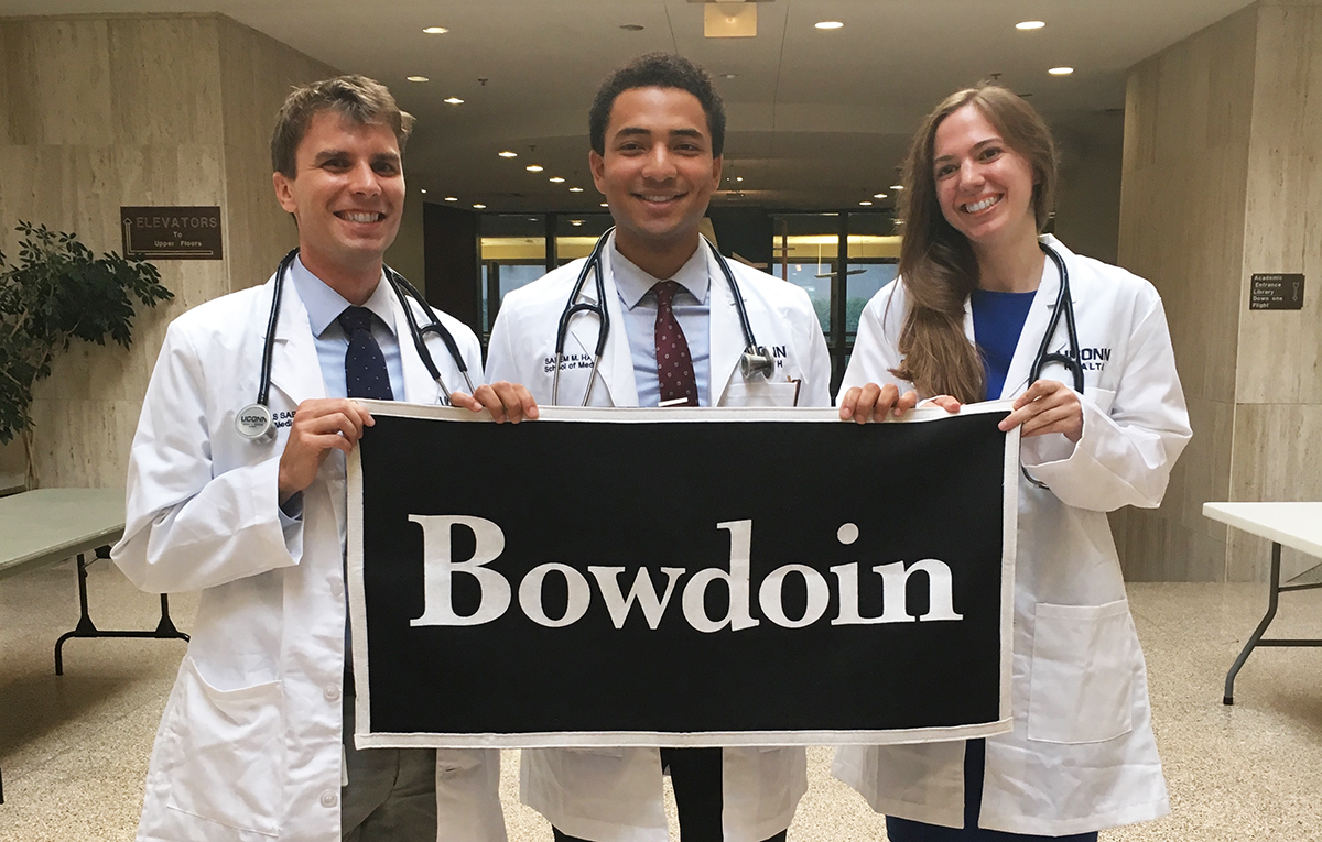 3 students in white medical coats hold up Bowdoin Sign 