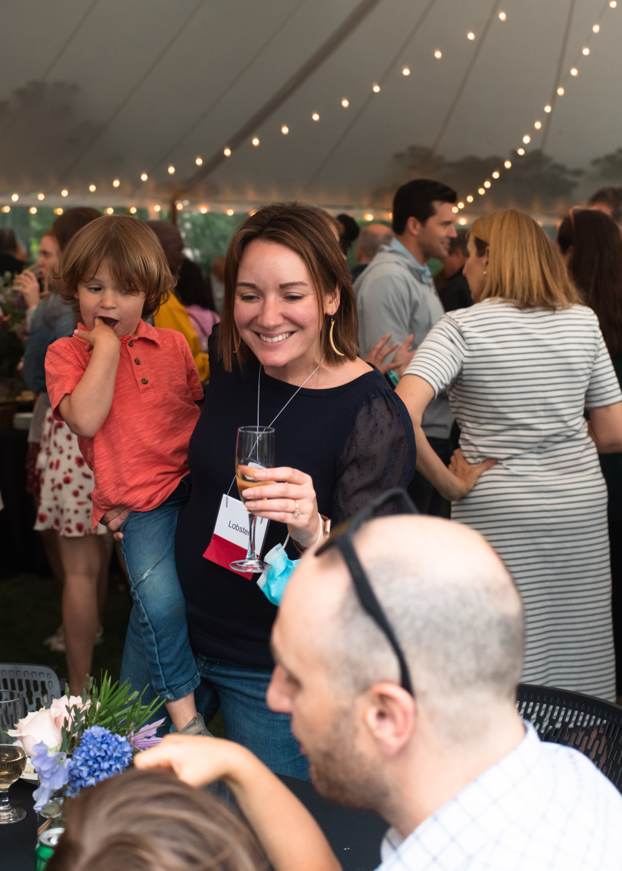 A woman smiling and looking down at a table in the event tent as she holds a young child to her hip.