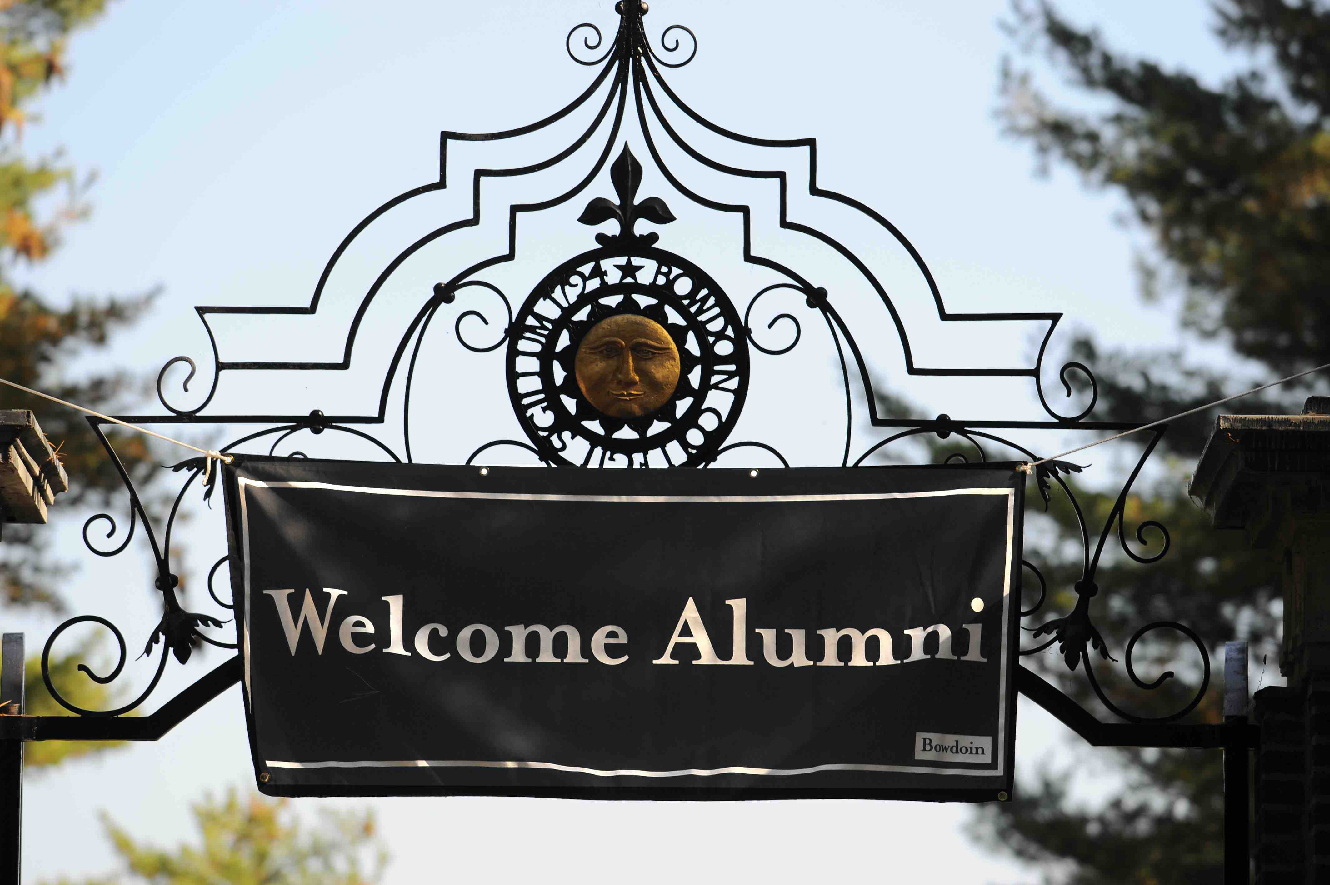 Image of brass gate with sun logo. Sign hangs from gate reading "Welcome Alumni." 