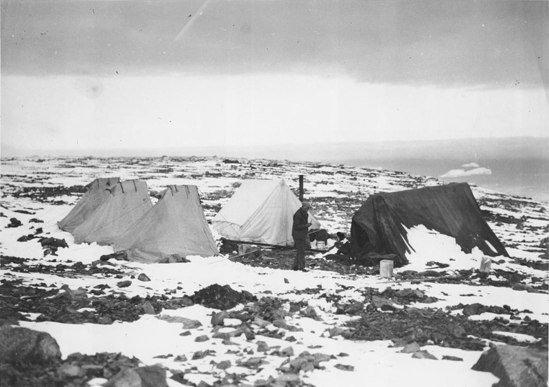 <p><b>Camp Scenes</b> </p>   We camped about 200 feet below the top of the Cape on a southern slope. The thirty days which were spent in camp will always be remembered as outstanding to each member of the construction gang. We went onto the mountain with very light equipment, three small tents with a square tent 9 by 12 feet….when the sun shone or when it rained, water would flow through the rocks over the ice underneath. Hence we had no trouble to get water, and at times it was only necessary to push aside a few stones inside the tent and dip it up. Jack Angel.<br /> <br />   Unknown photographer, Campsite, Cape York, Greenland, 1932. Ink jet print from black and white photograph. Robert Abram Bartlett papers, George J. Mitchell Department of Special Collections & Archives, Bowdoin College Library.  