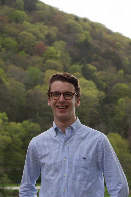 photo of Brandon Shuster smiling, wearing a light blue button down shirt, with a green forested hillside in the background
