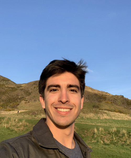 Photo of Ramiro Storni, smiling, wearing a gray t-shirt and brown jacket, with a blue sky and grassy hills in the background