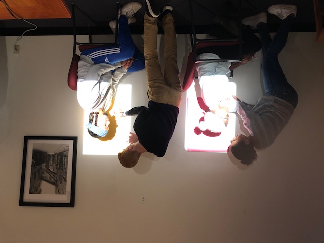 Students sit around Picture Study Room seminar table cutting out silhouettes, while another student is in background tracing another student's profile projected on wall. 