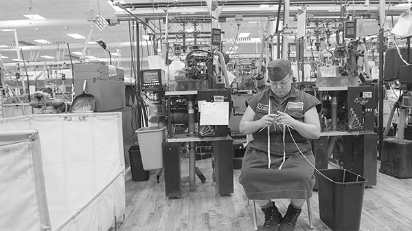 a black and white photo of a seated woman hand-knitting, in front of an industrial knitting machine