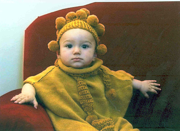 A photo of a baby in a knitted golden-colored outfit with pom-poms.