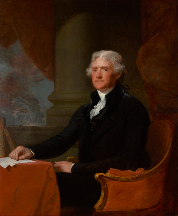 A gentleman with white hair seated in a chair in front of red curtains