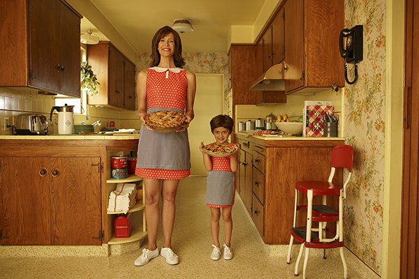 A woman and child in matching outfits of red polka dotted dresses with black and white checked aprons holding  pies in a 1950s looking kitchen