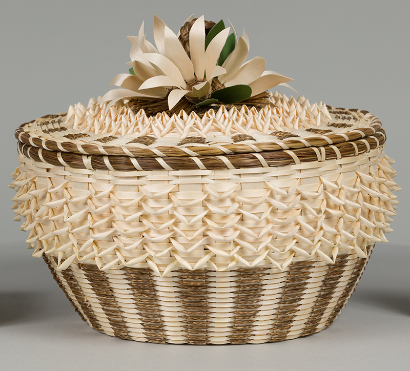 a beige and tan basket with a flower on the lid and fancy weaving