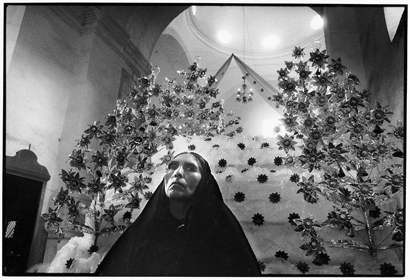 a black and white photo of a woman in a dark shawl on her head. She is in an room with shiny metal stars/objects suspended above her head.