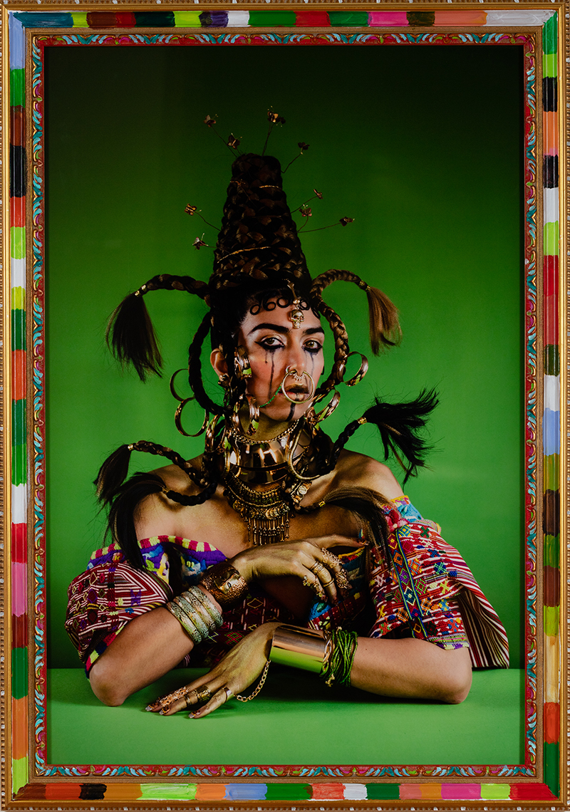 A color photograph of a figure with braided hair, jewlery, and rings tassels, jewelrey  agains a green background