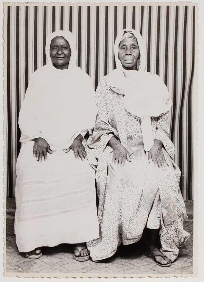 a black and white photo of two seated people, in light colored garments and head coverings,in front of a striped background
