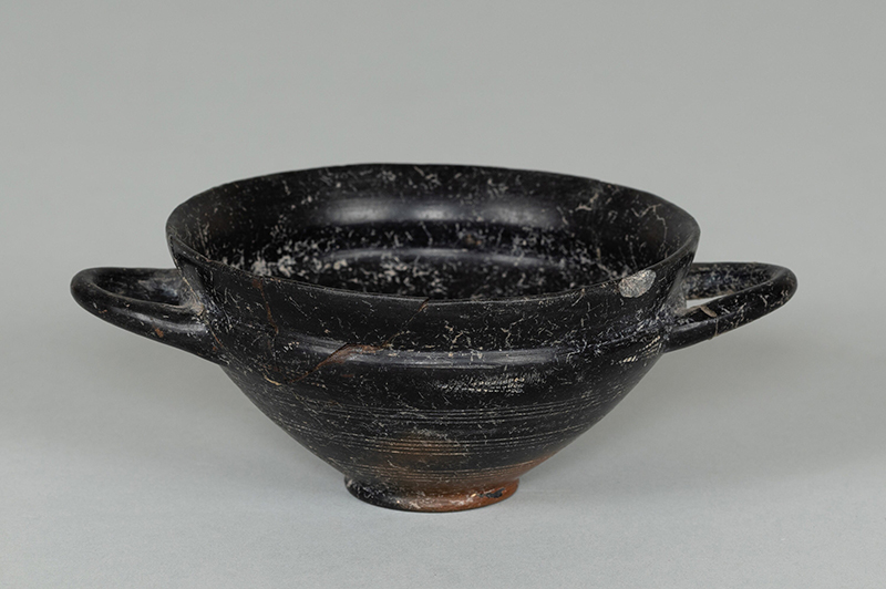 An ancient, black, drinking cup has a broad, shallow bowl with two handles atop a pedestal base.  