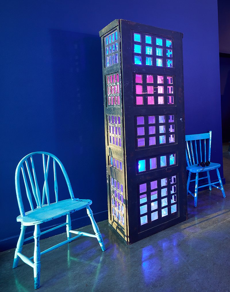 A rectangular sculpture with window like openings showing purple and pink lights, flanked by two chairs.