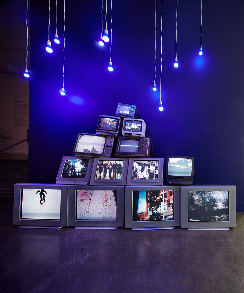 An assemblage sculpture of reclaimed televisions with videos playing under a group of light bulbs on long cords