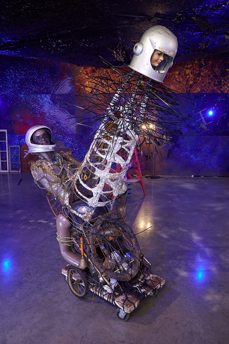 A sculpture made of found objects, showing two partial figures, with costume jewelry, wire, foil, all on wheels