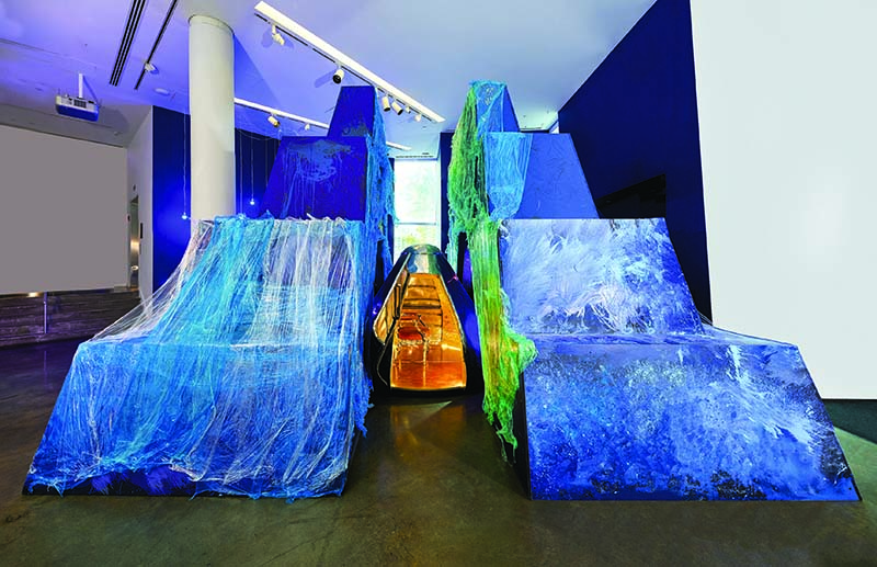 an art installation with two large blue cascading pieces of fabric on either side and a constructed object resembling a lunar capsule in the center.