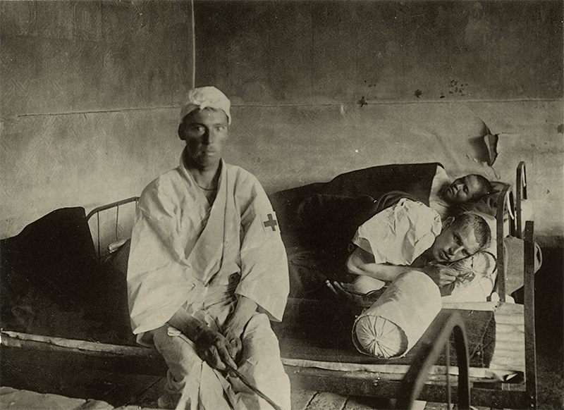 black and white photograph showing a person with a bandaged head sitting on a hospital bed occupied by two patientson a hos
