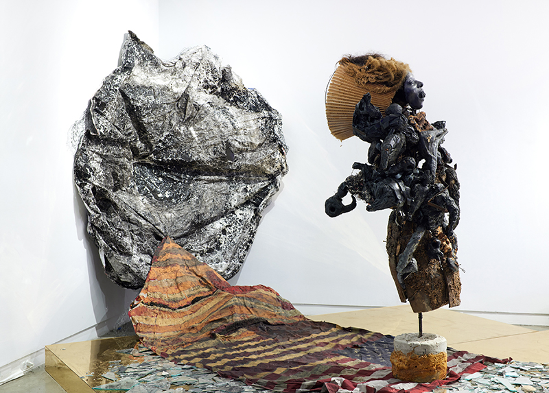 An assemblage sculpture with an organic form on the left, a fabric and broken glass cloth on the floor, and a  figure on the right