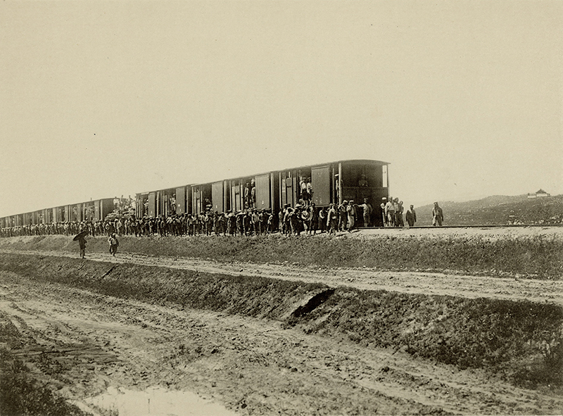 Sepia photo of a train in a barren country landscape, with many soldiers surrounding the train.