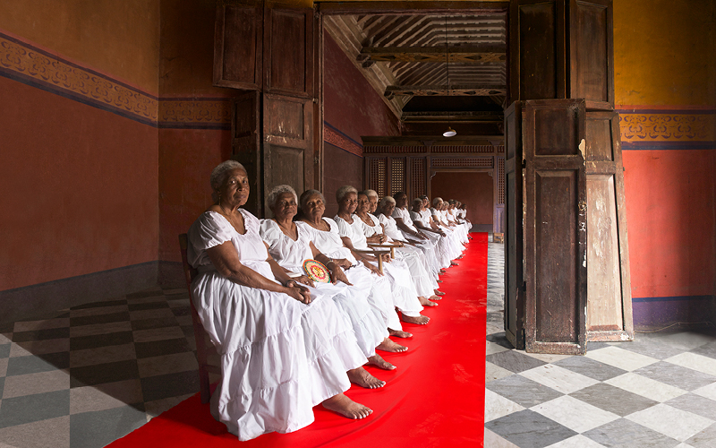 Women in white dress sit in a row on a red carpet facing the camera