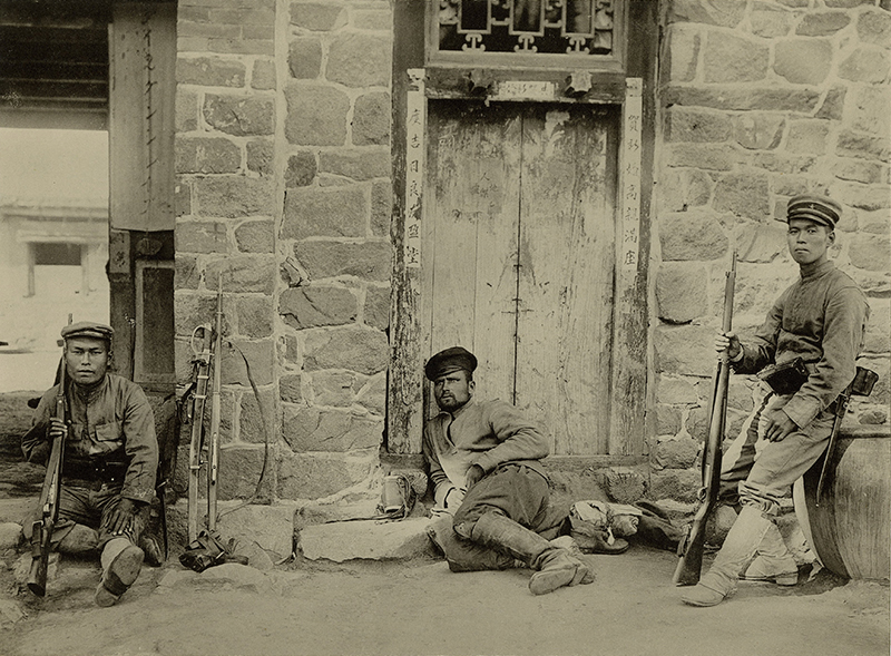 Sepia photograph showing three soldiers in front a stone building with a wooden door  Two people hold rifles, the third is half-lying down near the door.