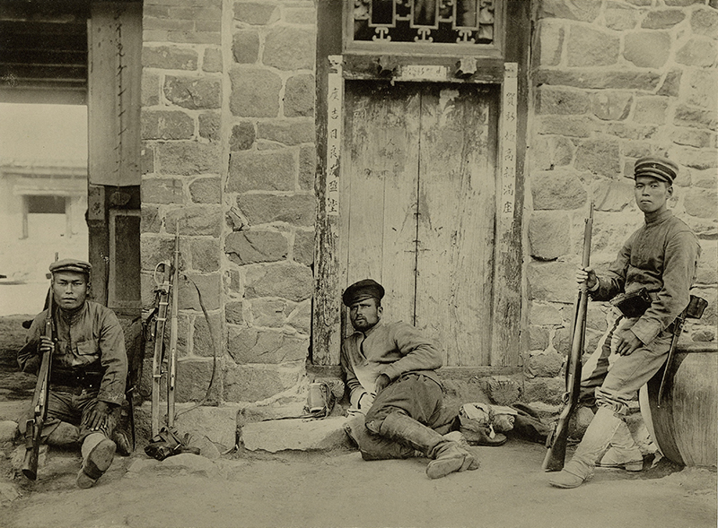 Sepia photograph showing three soldiers in front a stone building with a wooden door Two people hold rifles, the third is half-lying down near the door.