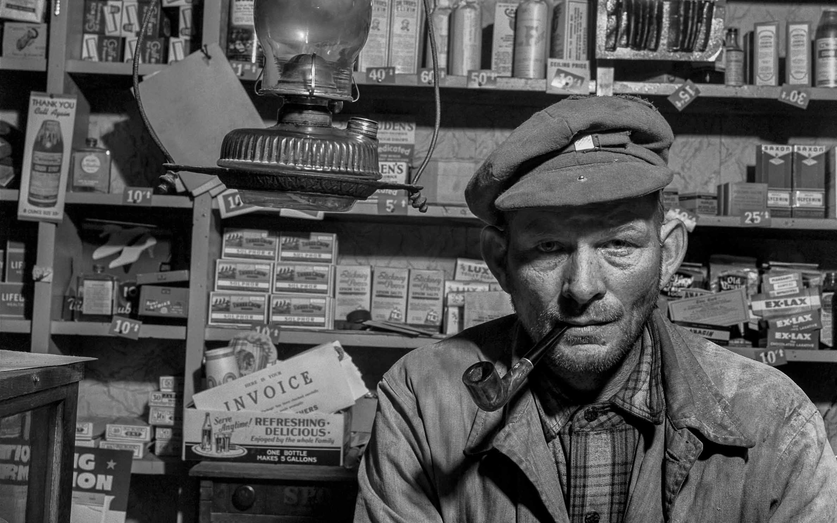 A detail of a black and white photograph shows a general store owner seated in front of his merchandise