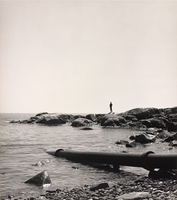 A black and white photograph shows a person fishing and large metal pipe leading into the ocean