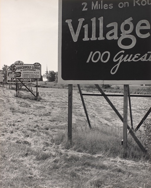A black and white photograph depicts roadside billboard advertisements