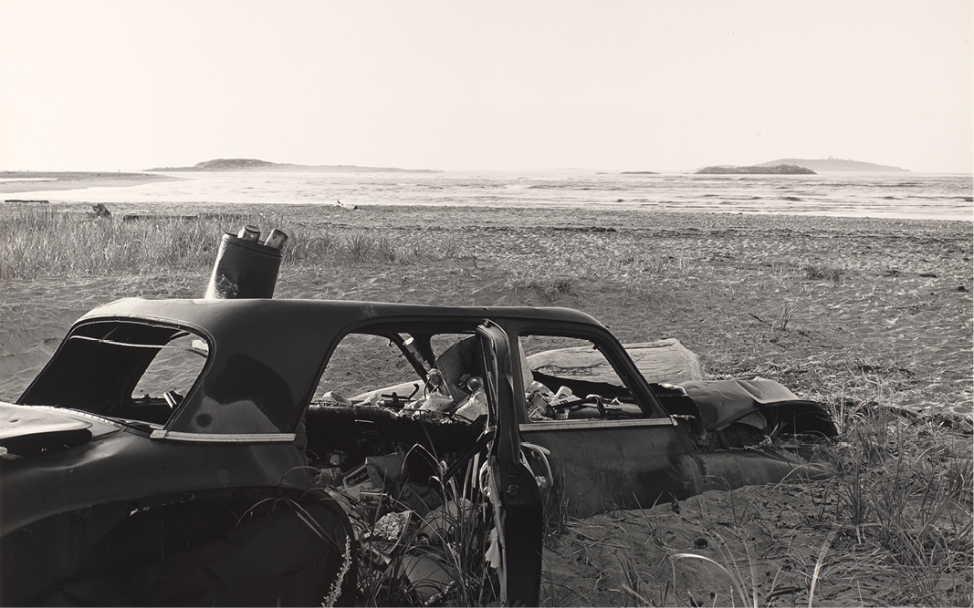 A detail of a black and white photograph shows a rusted shell of a car in a coastal landscape