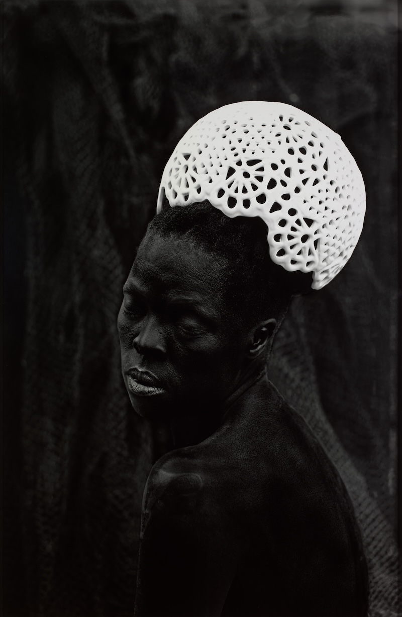 A detail of a black and white photograph of a figure with dark skin wearing a white lattice crown set against a dark background