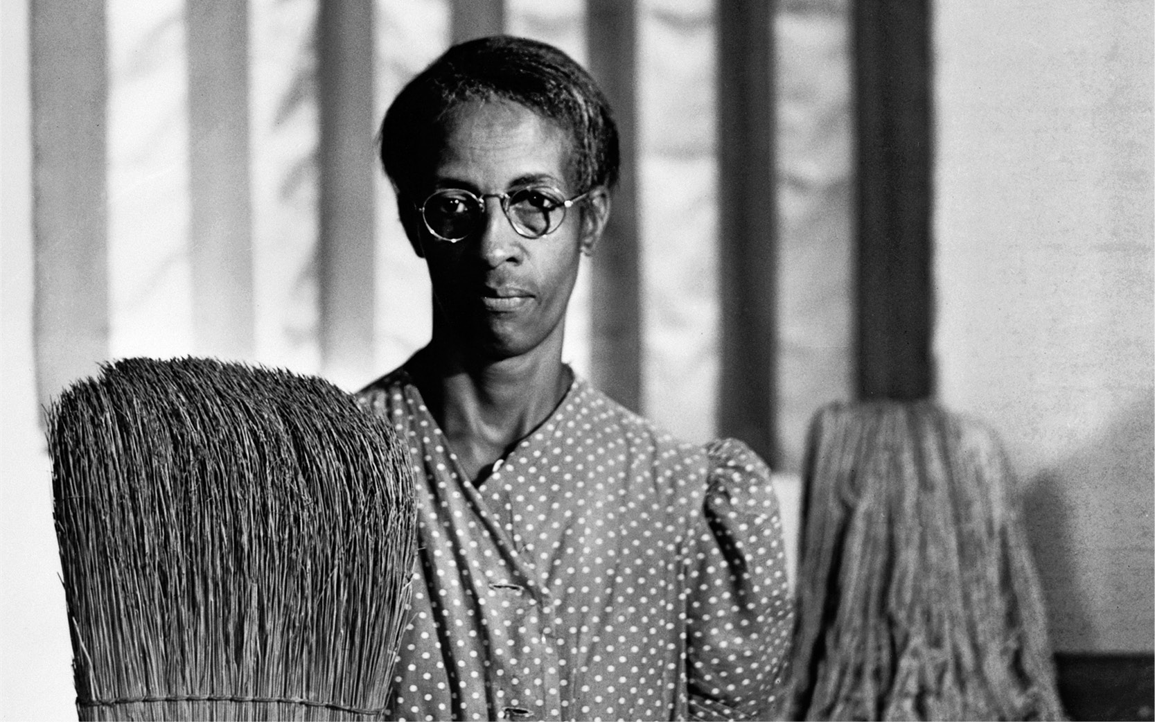A detail of a black and white photograph of a Black woman in a polka dot shirt holding a broom and mop in front of an American flag