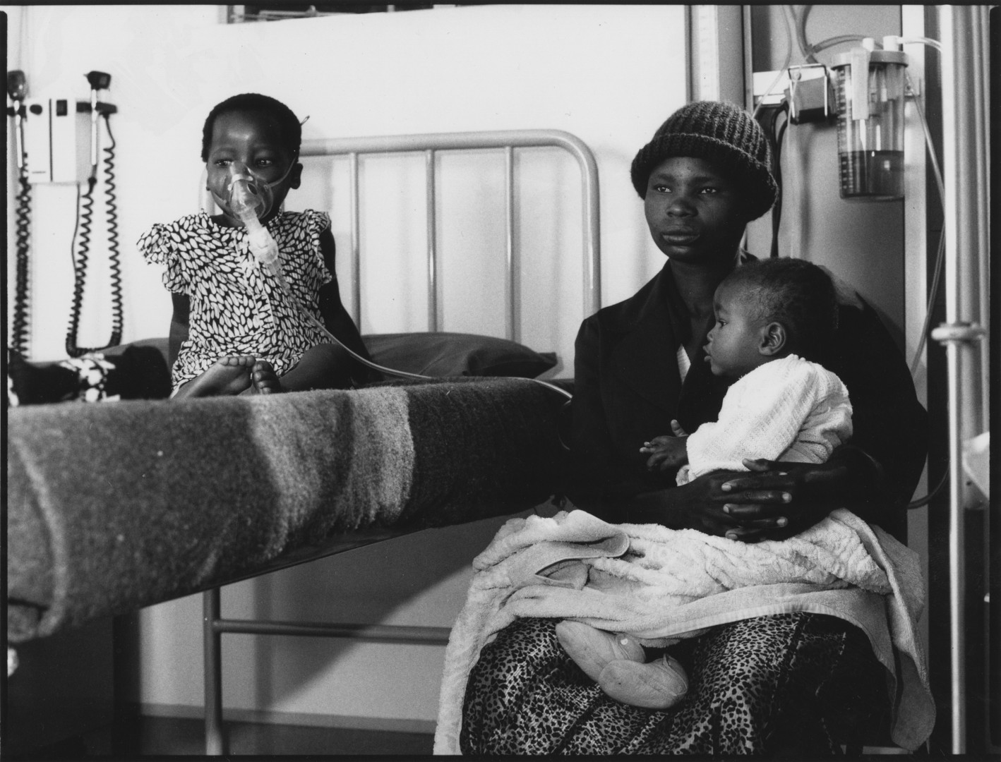 a woman sits with a child on her lap. to her left a child sits on a bed with an oxygen mask