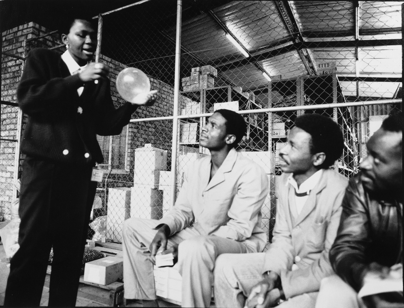 a person holds an inflated condom while three sitted men watch.