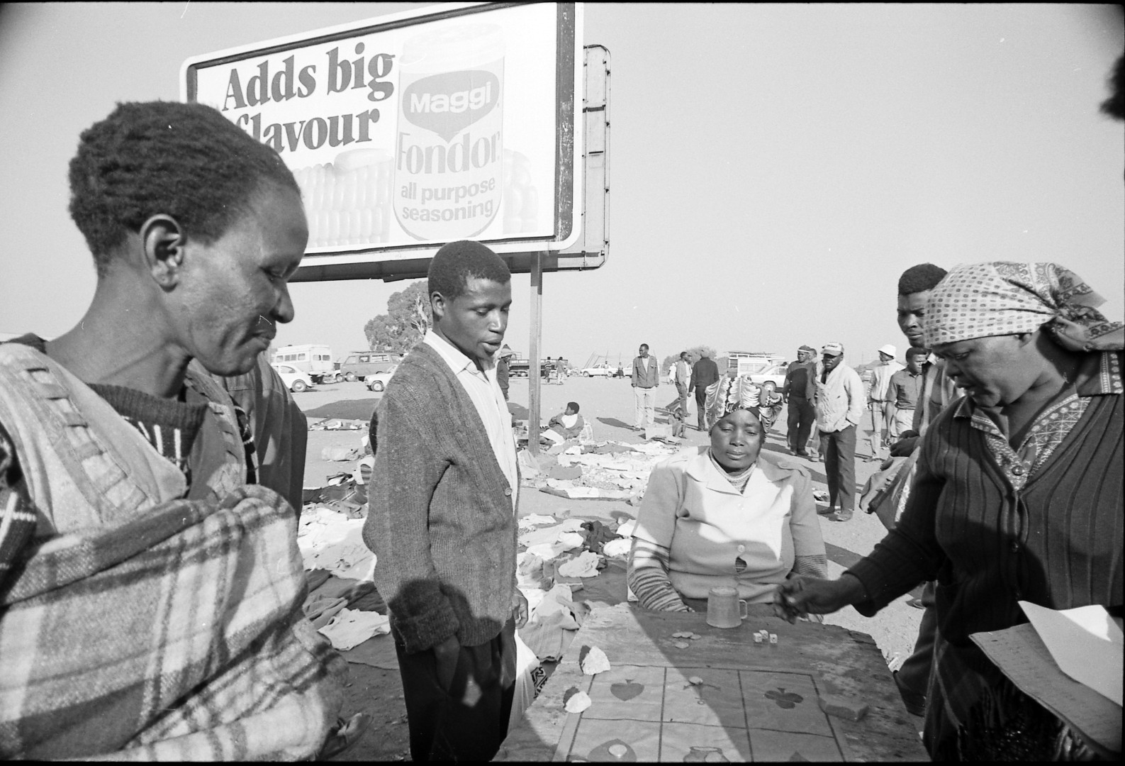In the foreground men and women gather around a table for gambling. In the background is a large billboard and more people and cars.