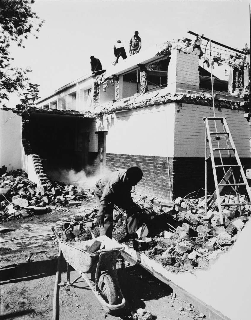 a man works among rubble with a wheelbarrow. in the background mean stand on a roof of a burned-out building.