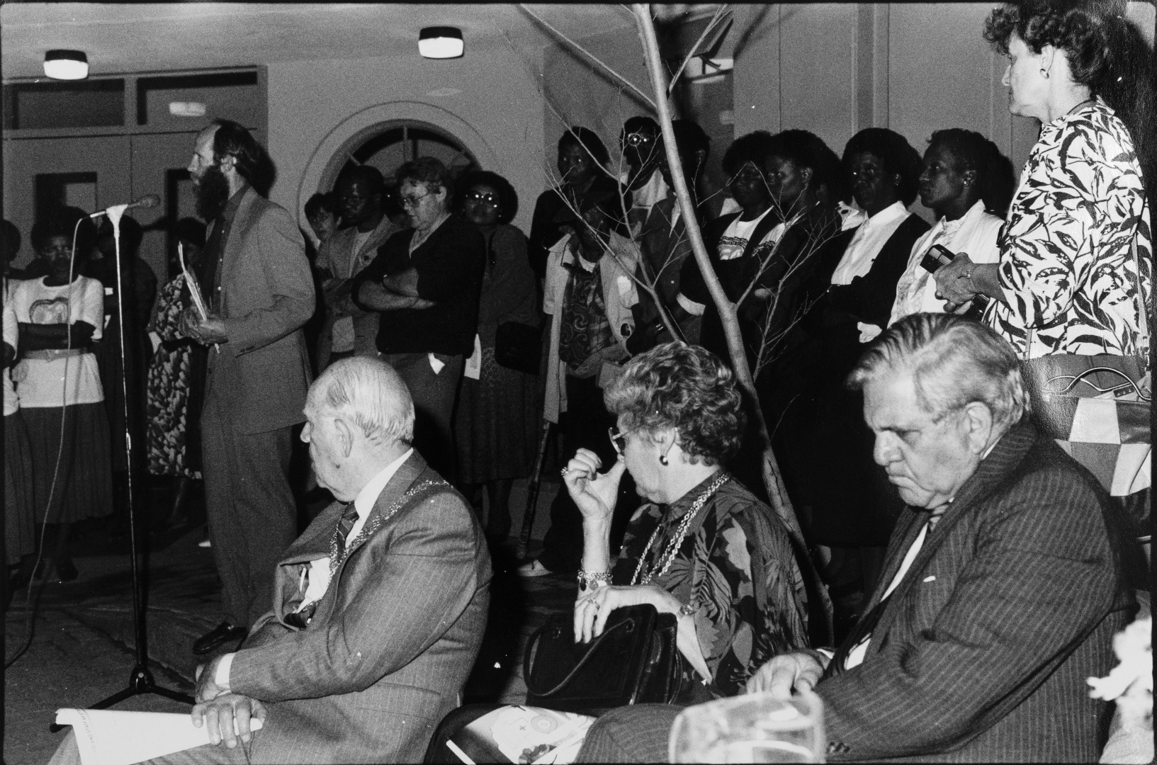 well-dressed people sit in the foreground. A man speaks into a microphone behind them while several people stand listening.