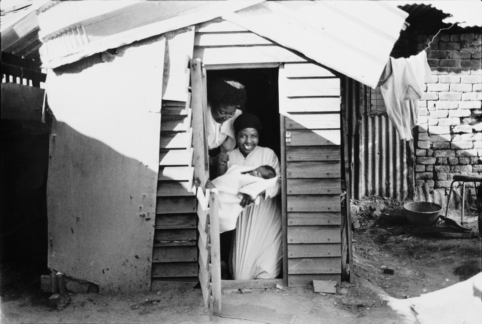 Two women in the doorway of a small shack. One holds a baby in her arms.