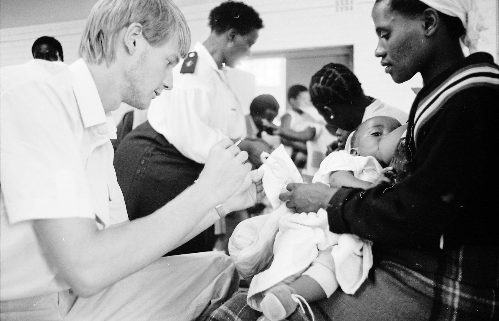 in the foreground a woman breastfeeds a baby as a man administers medicine.