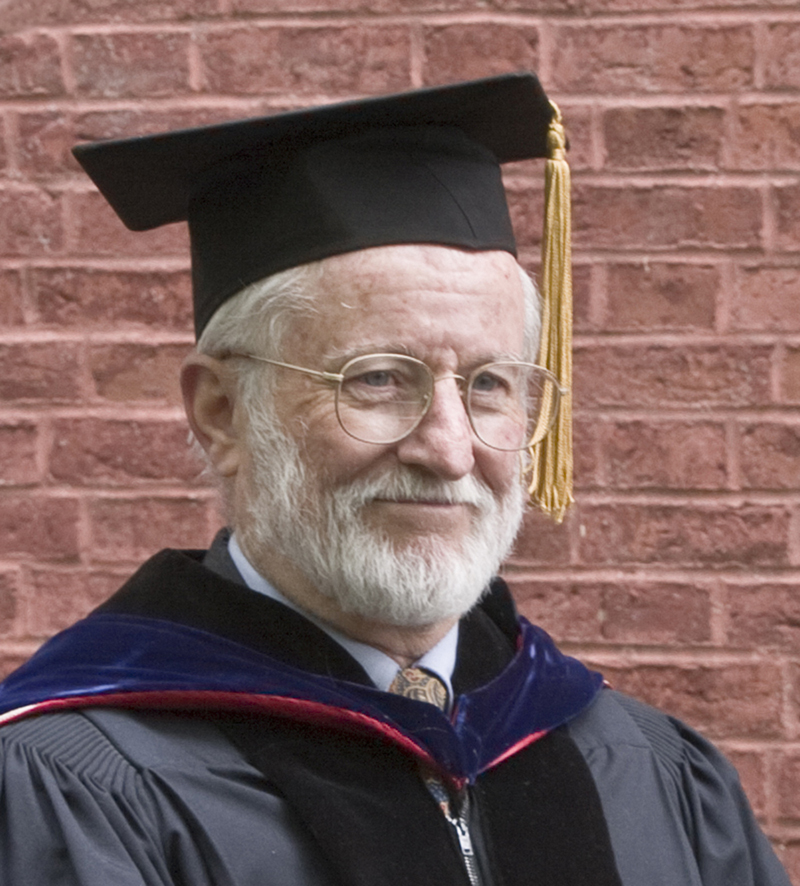 A photograph of a man with glasses in an academic cap, in front of a brick wall