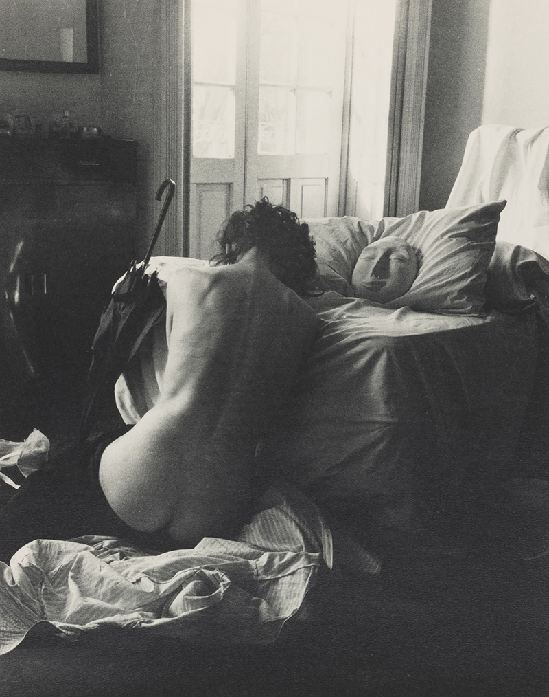 a black and white photograph showing a seated person's unclothed back, a bed and other drapery