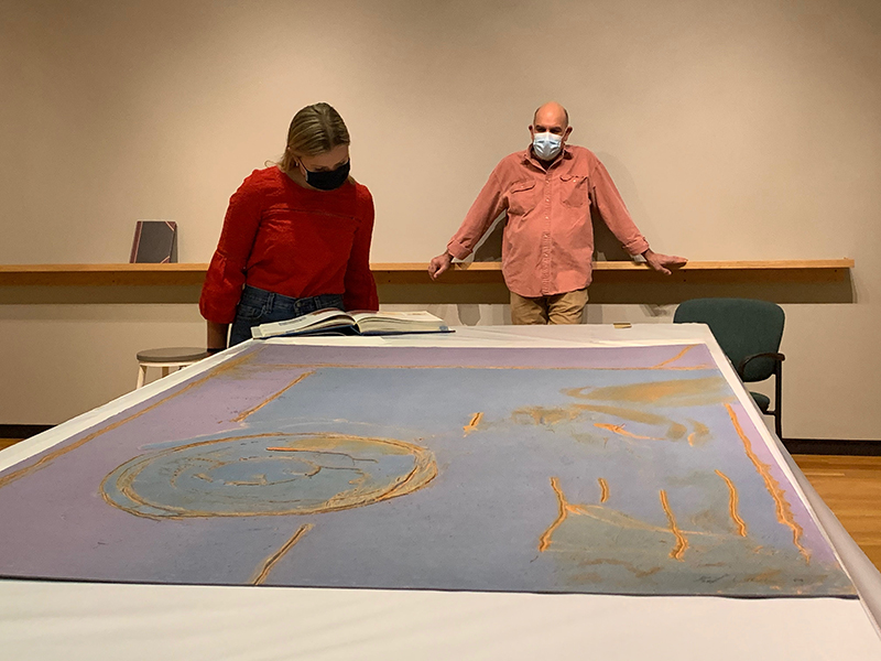 Two figures in a museum, one studying a work of art on a table