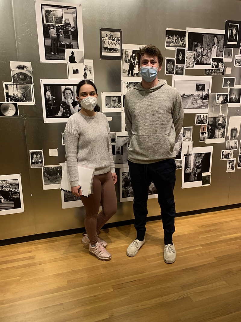 Two students standing in a gallery with a collage of photographs behind them