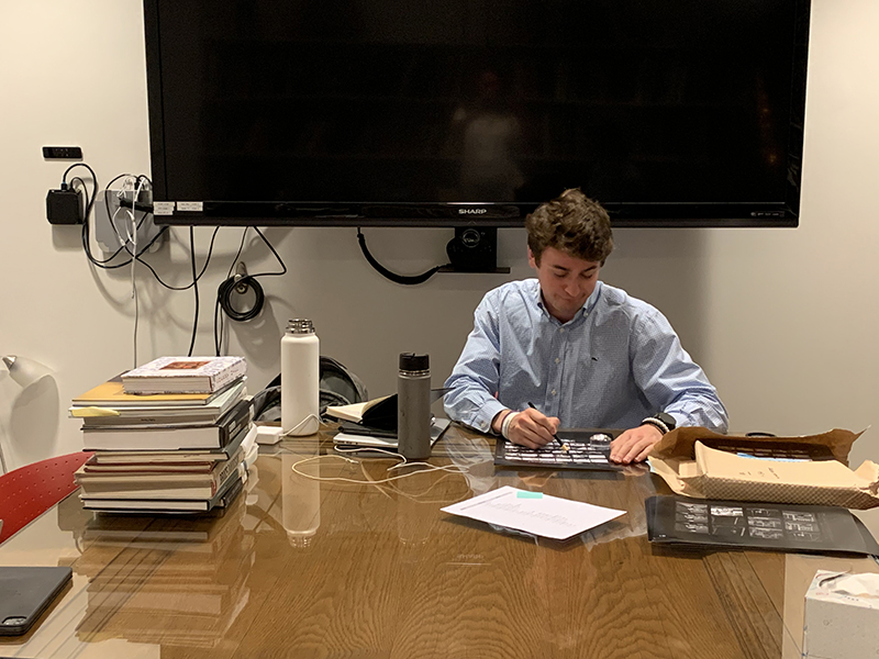 A person reading, seated at a table, with a pile of books and research materials