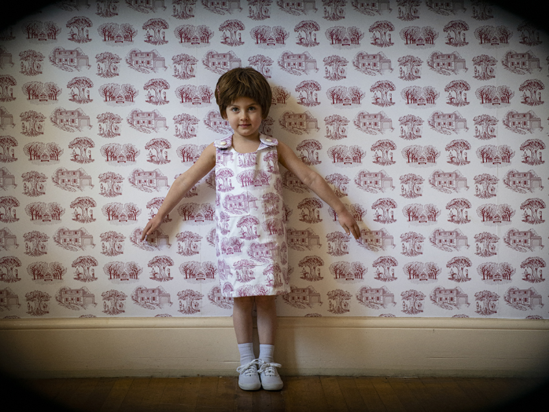 A young girl standing  in front of a wall-papered wall.  Her dress matches the wallpaper pattern.