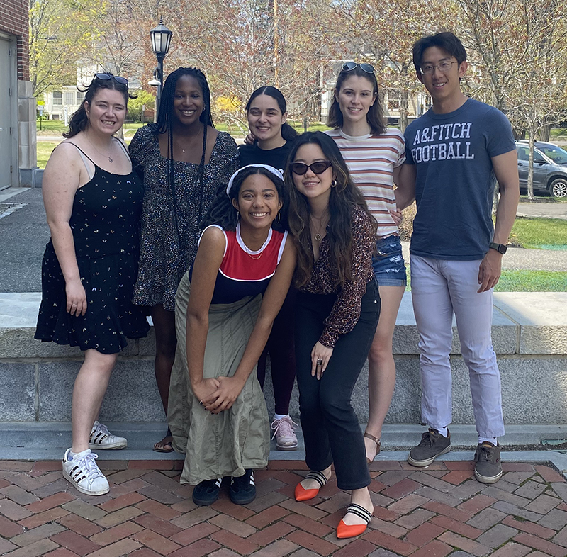 A group photo of seven Bowdoin students, outdoors standing on a brick surface with a blue sky behind them