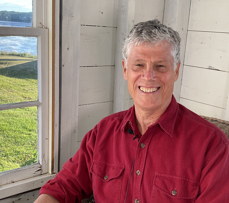 A smiling man in a red shirt in front of a wall of boards with a window to the left