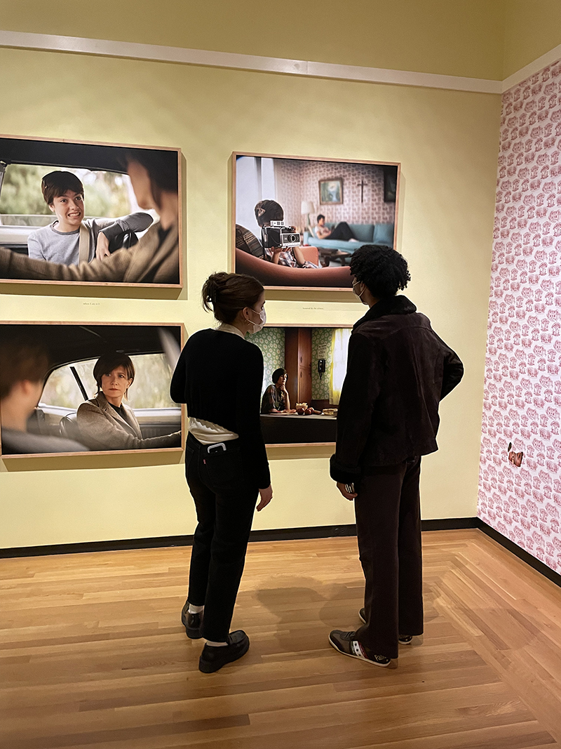 Two figures standing in front of a yellow gallery wall with four large photographs