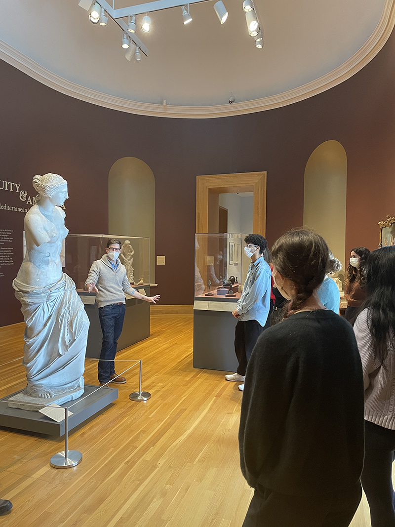 A group of people listening to a discussion in a gallery.  The speaker stands next to a large classical sculpture