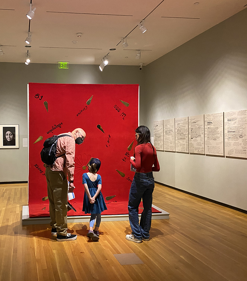 Three human figures standing in a gallery in front of a bright red rug